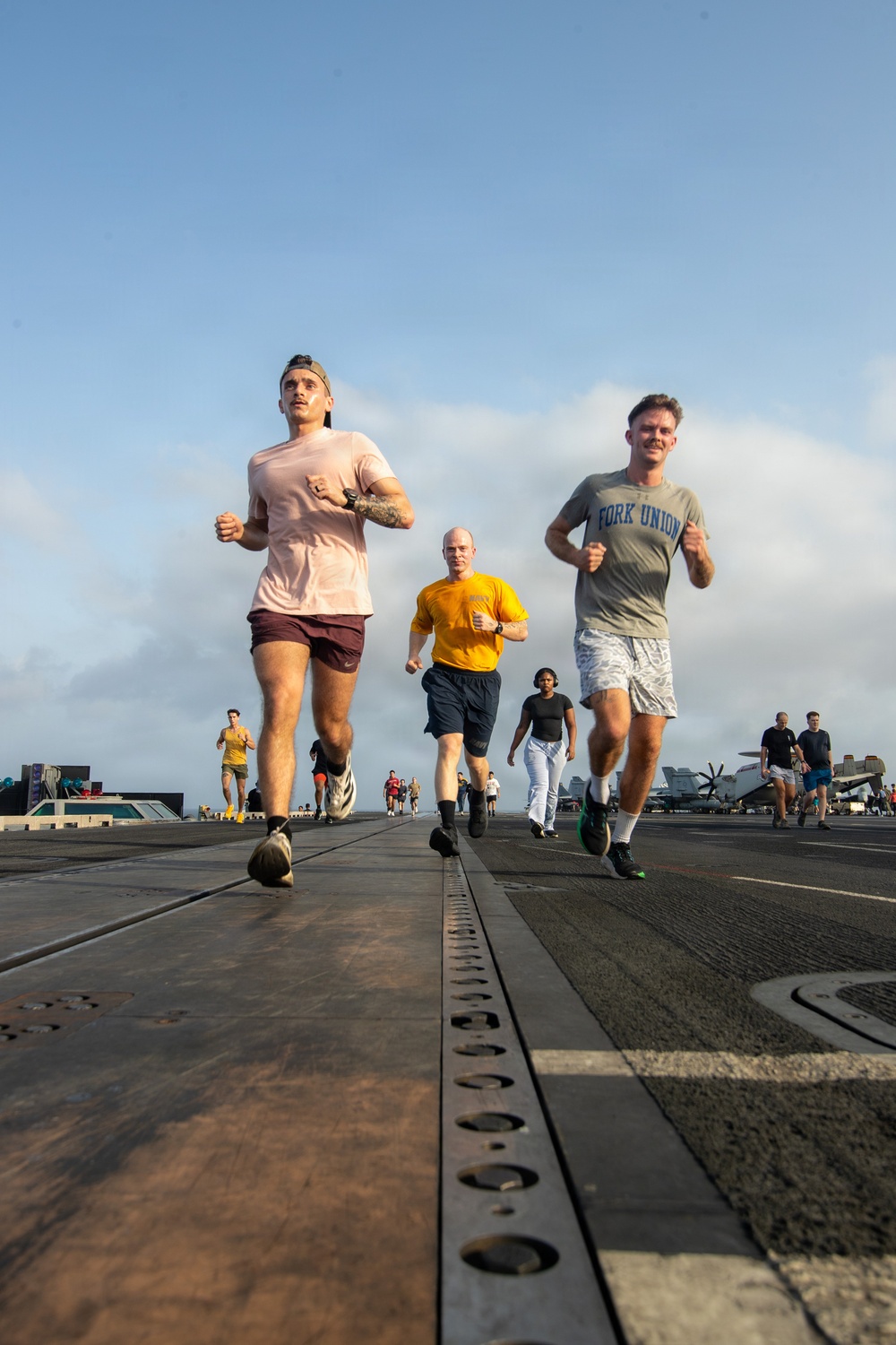 A group of military personnel are jogging on the flight deck of an aircraft carrier during what appears to be a physical training session. The low-angle photograph captures multiple runners in civilian athletic wear moving toward the camera, with aircraft and flight deck equipment visible in the background. The setting is clearly a naval vessel at sea under partly cloudy skies.
