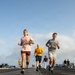 A group of military personnel are jogging on the flight deck of an aircraft carrier during what appears to be a physical training session. The low-angle photograph captures multiple runners in civilian athletic wear moving toward the camera, with aircraft and flight deck equipment visible in the background. The setting is clearly a naval vessel at sea under partly cloudy skies.