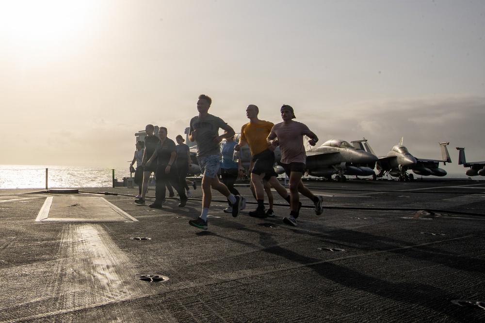 A group of military personnel are jogging on the flight deck of an aircraft carrier at what appears to be dawn or dusk, with the sun low on the horizon creating a backlit silhouette effect. Several jet aircraft, likely F/A-18 Hornets or similar carrier-based fighters, are parked on the deck in the background. The scene depicts a physical fitness exercise routine conducted aboard a naval vessel at sea.