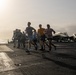A group of military personnel are jogging on the flight deck of an aircraft carrier at what appears to be dawn or dusk, with the sun low on the horizon creating a backlit silhouette effect. Several jet aircraft, likely F/A-18 Hornets or similar carrier-based fighters, are parked on the deck in the background. The scene depicts a physical fitness exercise routine conducted aboard a naval vessel at sea.