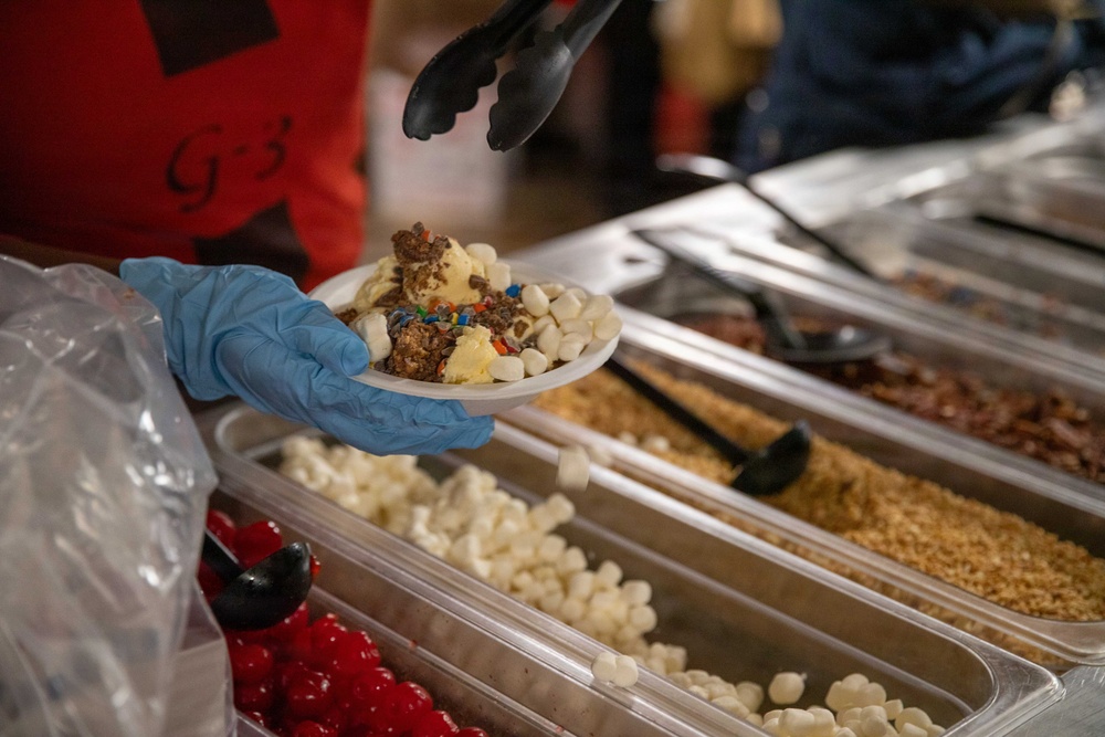 A food service worker wearing a blue latex glove and red uniform holds a white plate topped with ice cream scoops and multiple toppings including mini marshmallows, sprinkles, and granola. The scene is set at a dessert topping bar with multiple stainless steel trays containing various toppings such as mini marshmallows, graham cracker crumbs, maraschino cherries, and chopped nuts. The worker is using black tongs to add or arrange toppings on the dessert.