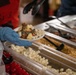 A food service worker wearing a blue latex glove and red uniform holds a white plate topped with ice cream scoops and multiple toppings including mini marshmallows, sprinkles, and granola. The scene is set at a dessert topping bar with multiple stainless steel trays containing various toppings such as mini marshmallows, graham cracker crumbs, maraschino cherries, and chopped nuts. The worker is using black tongs to add or arrange toppings on the dessert.