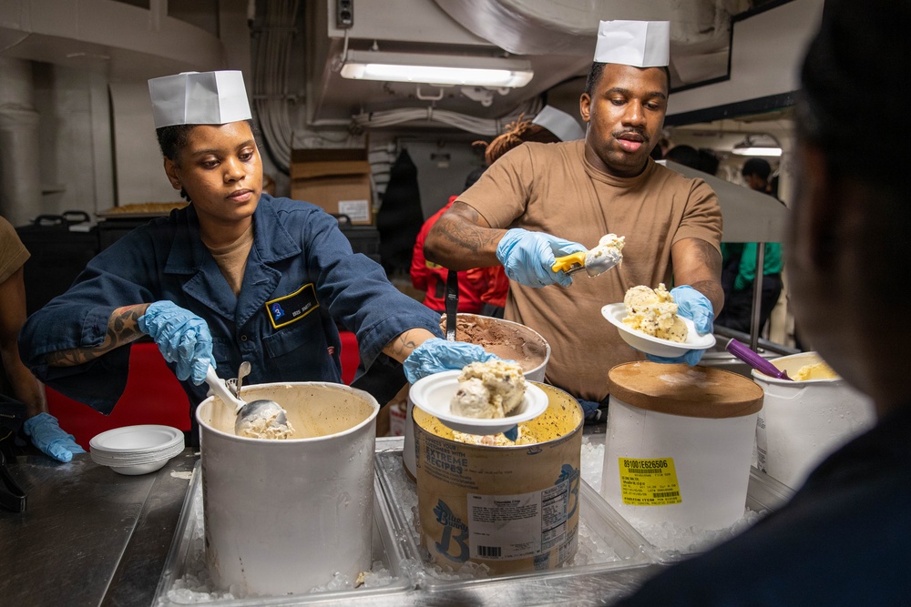 Two individuals wearing paper chef hats and blue latex gloves are serving ice cream from large commercial containers packed in ice. The setting appears to be a military galley or mess hall, indicated by the naval uniform worn by the woman on the left and the industrial kitchen environment. Multiple flavors of ice cream are being scooped onto plates for service.