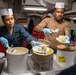 Two individuals wearing paper chef hats and blue latex gloves are serving ice cream from large commercial containers packed in ice. The setting appears to be a military galley or mess hall, indicated by the naval uniform worn by the woman on the left and the industrial kitchen environment. Multiple flavors of ice cream are being scooped onto plates for service.