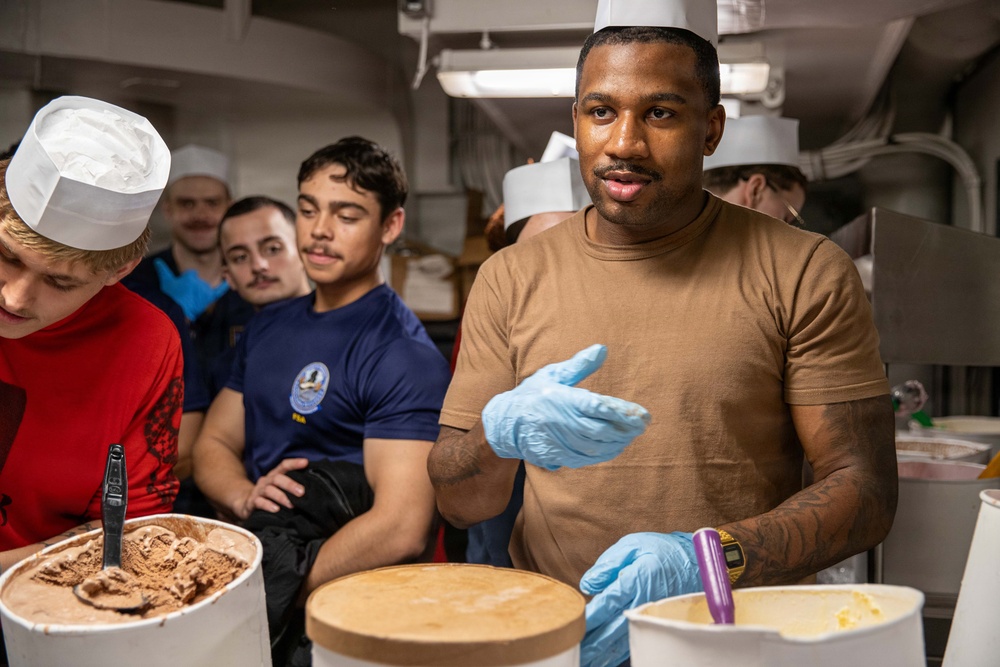A group of individuals in a kitchen or galley setting, with one person in a tan shirt and blue latex gloves appearing to instruct or demonstrate something related to food preparation. Several large containers of what appear to be ice cream or frozen desserts are visible in the foreground. Some participants are wearing white chef's hats, suggesting a culinary training or food service context.