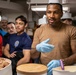 A group of individuals in a kitchen or galley setting, with one person in a tan shirt and blue latex gloves appearing to instruct or demonstrate something related to food preparation. Several large containers of what appear to be ice cream or frozen desserts are visible in the foreground. Some participants are wearing white chef's hats, suggesting a culinary training or food service context.