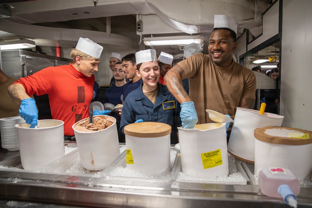 A group of individuals wearing white paper chef hats and blue latex gloves are serving ice cream from large white containers packed in ice, likely in a military galley or mess hall setting. The scene appears festive and cheerful, with participants smiling and laughing. The setting, uniforms, and 'G-3' markings suggest a U.S. Navy shipboard environment.