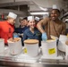A group of individuals wearing white paper chef hats and blue latex gloves are serving ice cream from large white containers packed in ice, likely in a military galley or mess hall setting. The scene appears festive and cheerful, with participants smiling and laughing. The setting, uniforms, and 'G-3' markings suggest a U.S. Navy shipboard environment.