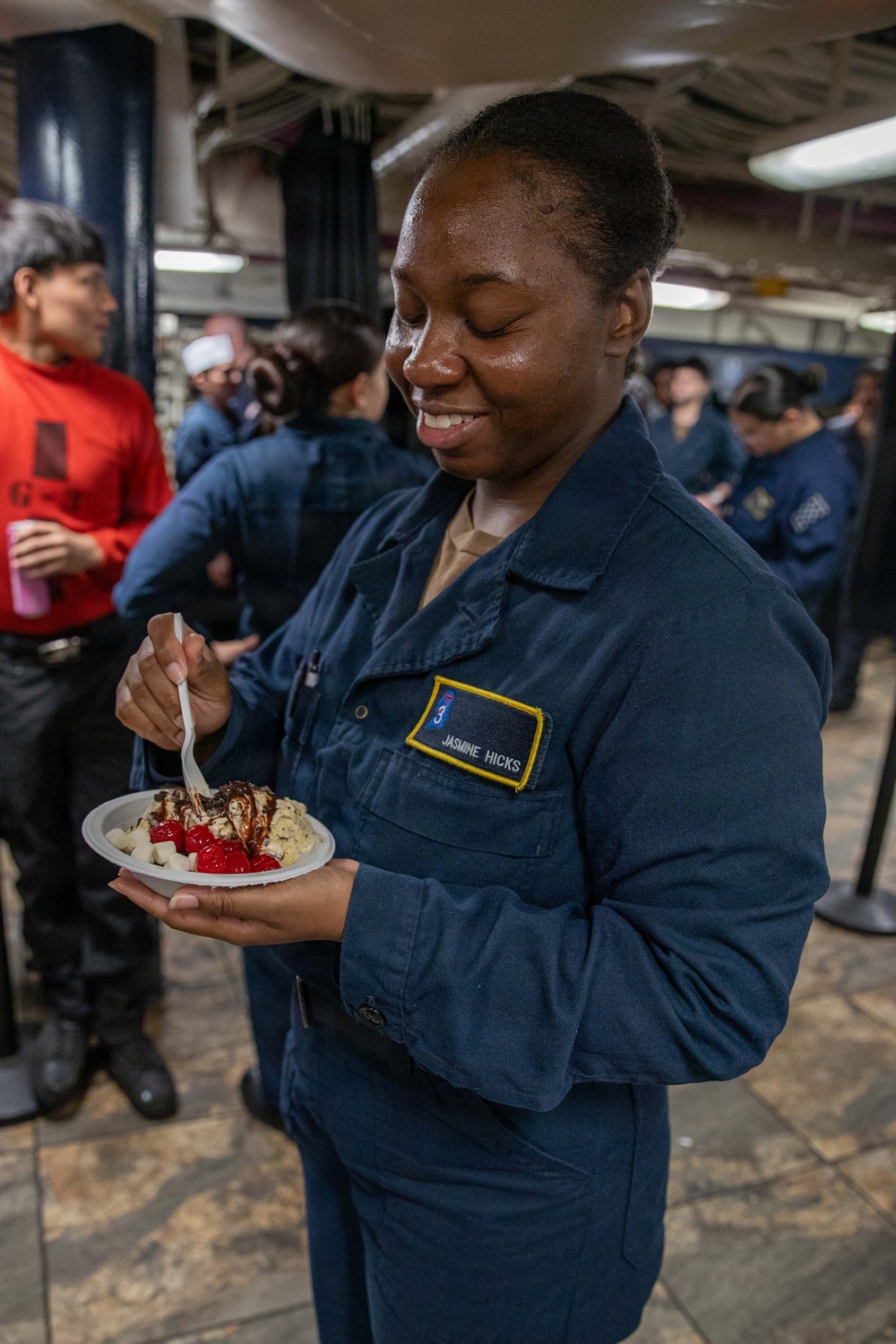 A U.S. Navy sailor named Jasmine Hicks, wearing blue Navy coveralls (NWU Type III), is smiling with eyes closed while holding and eating a dessert bowl containing what appears to be ice cream with cherries and chocolate sauce. She is standing in what appears to be a shipboard common area or mess deck, surrounded by other personnel in the background. The scene suggests a morale event or special meal occasion aboard a naval vessel.