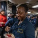 A U.S. Navy sailor named Jasmine Hicks, wearing blue Navy coveralls (NWU Type III), is smiling with eyes closed while holding and eating a dessert bowl containing what appears to be ice cream with cherries and chocolate sauce. She is standing in what appears to be a shipboard common area or mess deck, surrounded by other personnel in the background. The scene suggests a morale event or special meal occasion aboard a naval vessel.
