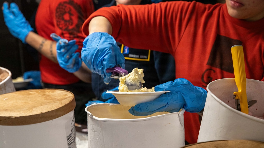 Workers wearing red shirts and blue latex gloves are serving food, appearing to scoop a creamy or starchy substance onto small white paper plates. The scene suggests a food service or community event setting with multiple large white containers visible. The close-up perspective emphasizes the food handling and serving activity.