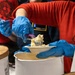 Workers wearing red shirts and blue latex gloves are serving food, appearing to scoop a creamy or starchy substance onto small white paper plates. The scene suggests a food service or community event setting with multiple large white containers visible. The close-up perspective emphasizes the food handling and serving activity.