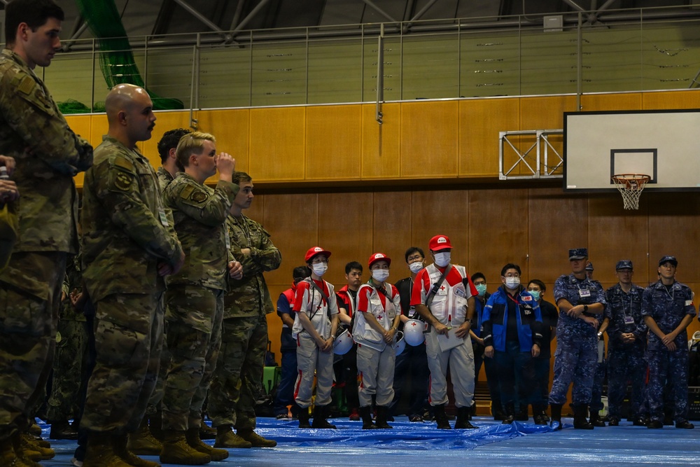 The image shows a multinational military and humanitarian coordination gathering inside a gymnasium or indoor sports facility. Personnel in U.S. Army camouflage uniforms face a group of Red Cross medical workers wearing red caps, white uniforms with red cross insignia, and face masks, alongside individuals in blue uniforms and naval personnel. The setting appears to be a joint disaster relief or humanitarian assistance exercise or briefing.