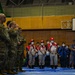 The image shows a multinational military and humanitarian coordination gathering inside a gymnasium or indoor sports facility. Personnel in U.S. Army camouflage uniforms face a group of Red Cross medical workers wearing red caps, white uniforms with red cross insignia, and face masks, alongside individuals in blue uniforms and naval personnel. The setting appears to be a joint disaster relief or humanitarian assistance exercise or briefing.
