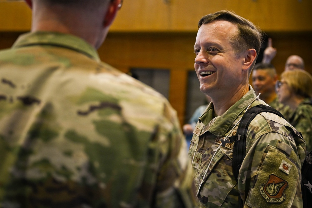 Two military personnel in OCP (Operational Camouflage Pattern) uniforms are engaged in conversation in what appears to be an indoor military facility. The soldier facing the camera is smiling broadly, suggesting a positive or informal interaction. Additional military and civilian personnel are visible in the background.