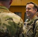 Two military personnel in OCP (Operational Camouflage Pattern) uniforms are engaged in conversation in what appears to be an indoor military facility. The soldier facing the camera is smiling broadly, suggesting a positive or informal interaction. Additional military and civilian personnel are visible in the background.