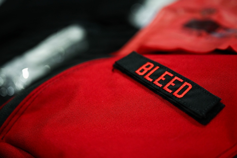 A close-up photograph of a red fabric item, likely a bag or tactical vest, featuring a black embroidered patch with the word 'BLEED' in red lettering. The image is shot with a shallow depth of field, keeping the patch in sharp focus while the background is blurred. Additional red fabric items and a metallic object are partially visible in the background.