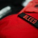 A close-up photograph of a red fabric item, likely a bag or tactical vest, featuring a black embroidered patch with the word 'BLEED' in red lettering. The image is shot with a shallow depth of field, keeping the patch in sharp focus while the background is blurred. Additional red fabric items and a metallic object are partially visible in the background.