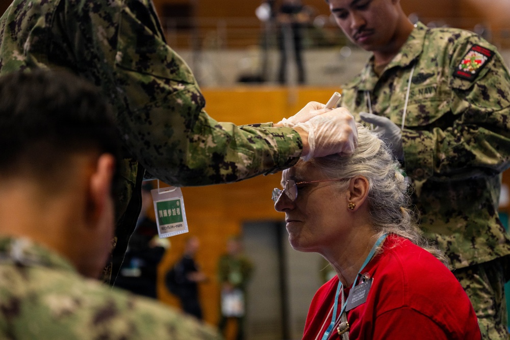 A U.S. Navy service member wearing latex gloves appears to be conducting a medical examination or applying treatment to an elderly woman's forehead or eye area. The setting appears to be an indoor facility, possibly during a humanitarian assistance or medical outreach event. Japanese text is visible on a badge/lanyard, suggesting this may be taking place in Japan.