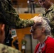 A U.S. Navy service member wearing latex gloves appears to be conducting a medical examination or applying treatment to an elderly woman's forehead or eye area. The setting appears to be an indoor facility, possibly during a humanitarian assistance or medical outreach event. Japanese text is visible on a badge/lanyard, suggesting this may be taking place in Japan.