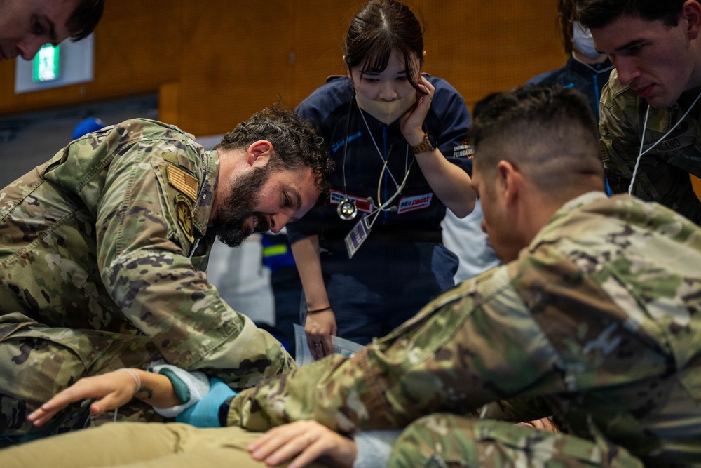 The image shows a group of military personnel in camouflage uniforms working alongside a civilian medical professional in navy scrubs during what appears to be a medical training exercise or joint exercise. A bearded soldier in OCP/MultiCam uniform is performing a hands-on medical procedure or assessment on a patient or mannequin on the floor. The setting appears to be an indoor facility, likely a training room or gymnasium.
