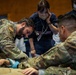 The image shows a group of military personnel in camouflage uniforms working alongside a civilian medical professional in navy scrubs during what appears to be a medical training exercise or joint exercise. A bearded soldier in OCP/MultiCam uniform is performing a hands-on medical procedure or assessment on a patient or mannequin on the floor. The setting appears to be an indoor facility, likely a training room or gymnasium.