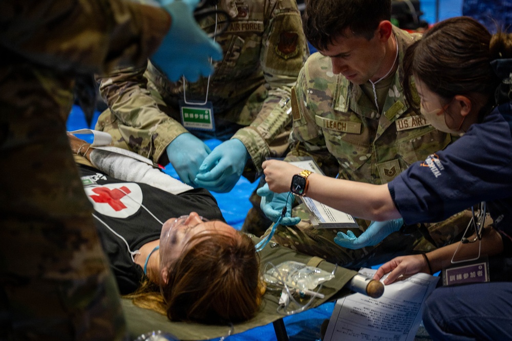 U.S. Air Force personnel are conducting a medical training exercise or disaster response drill, treating a simulated casualty lying on a stretcher. Personnel identified by name tape 'LEACH' and branch insignia 'U.S. AIR FORCE' are wearing MultiCam uniforms and blue medical gloves while attending to the patient. The presence of Chinese-language signage and badges suggests this is a joint or multinational exercise, likely held in Japan or another East Asian location.