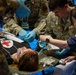 U.S. Air Force personnel are conducting a medical training exercise or disaster response drill, treating a simulated casualty lying on a stretcher. Personnel identified by name tape 'LEACH' and branch insignia 'U.S. AIR FORCE' are wearing MultiCam uniforms and blue medical gloves while attending to the patient. The presence of Chinese-language signage and badges suggests this is a joint or multinational exercise, likely held in Japan or another East Asian location.