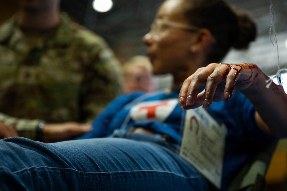 A woman in a blue jacket and jeans appears to be a simulated casualty in what looks like a military medical training exercise. Her hand shows simulated blood/moulage makeup, and she has a triage tag attached to her clothing. A person in military camouflage uniform is visible in the background.