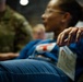 A woman in a blue jacket and jeans appears to be a simulated casualty in what looks like a military medical training exercise. Her hand shows simulated blood/moulage makeup, and she has a triage tag attached to her clothing. A person in military camouflage uniform is visible in the background.