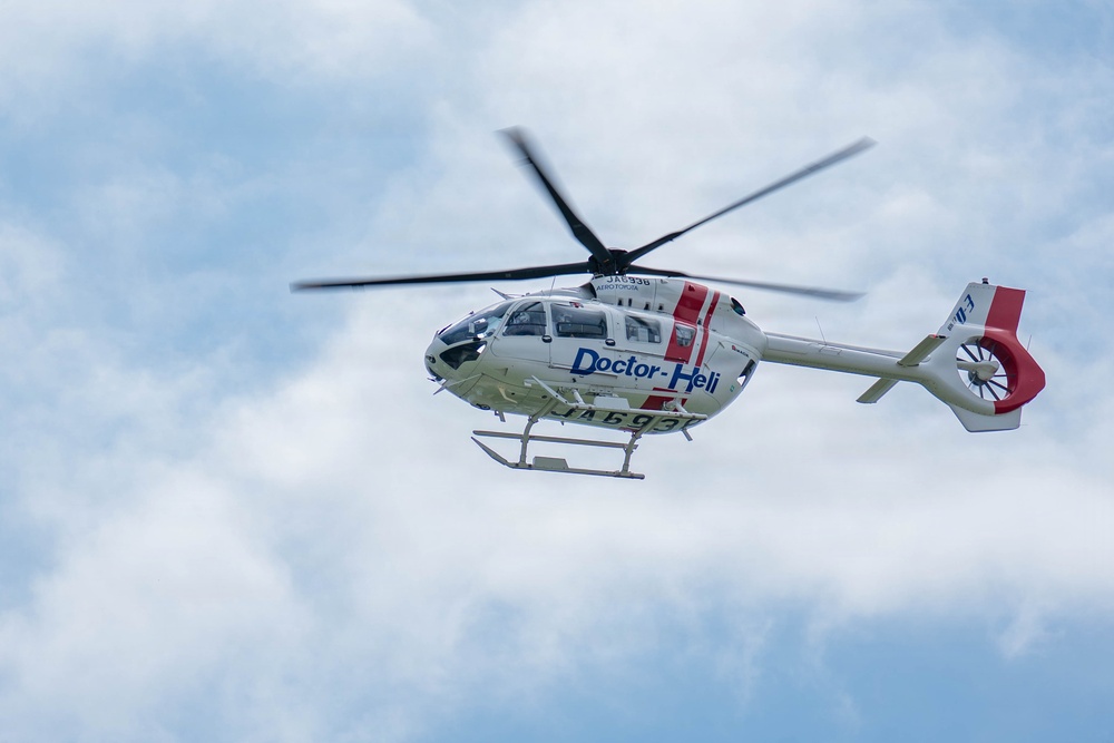 A white medical helicopter labeled 'Doctor-Heli' is captured in flight against a partly cloudy sky. The aircraft features red and blue markings and displays registration numbers on its fuselage. The helicopter appears to be a modern twin-engine model, likely used for emergency medical services.