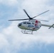 A white medical helicopter labeled 'Doctor-Heli' is captured in flight against a partly cloudy sky. The aircraft features red and blue markings and displays registration numbers on its fuselage. The helicopter appears to be a modern twin-engine model, likely used for emergency medical services.