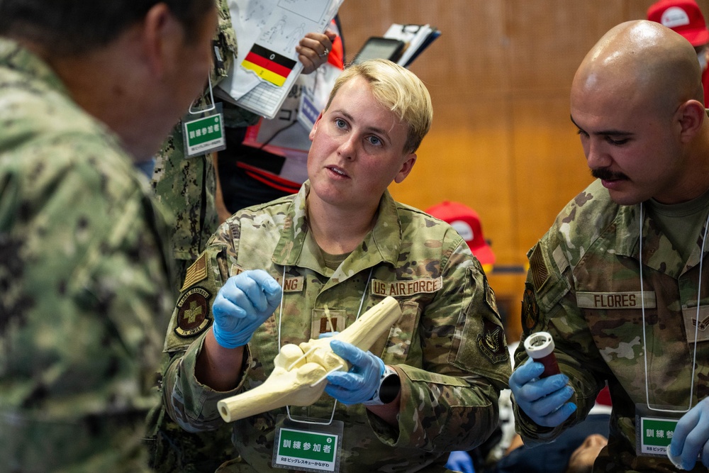 U.S. Air Force personnel are participating in a medical training exercise, likely in Japan based on the visible Japanese-language signage. A female Air Force member wearing blue medical gloves is holding and explaining an anatomical bone model, while a male colleague named Flores observes. The setting appears to be a multinational medical readiness training event.