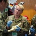 U.S. Air Force personnel are participating in a medical training exercise, likely in Japan based on the visible Japanese-language signage. A female Air Force member wearing blue medical gloves is holding and explaining an anatomical bone model, while a male colleague named Flores observes. The setting appears to be a multinational medical readiness training event.