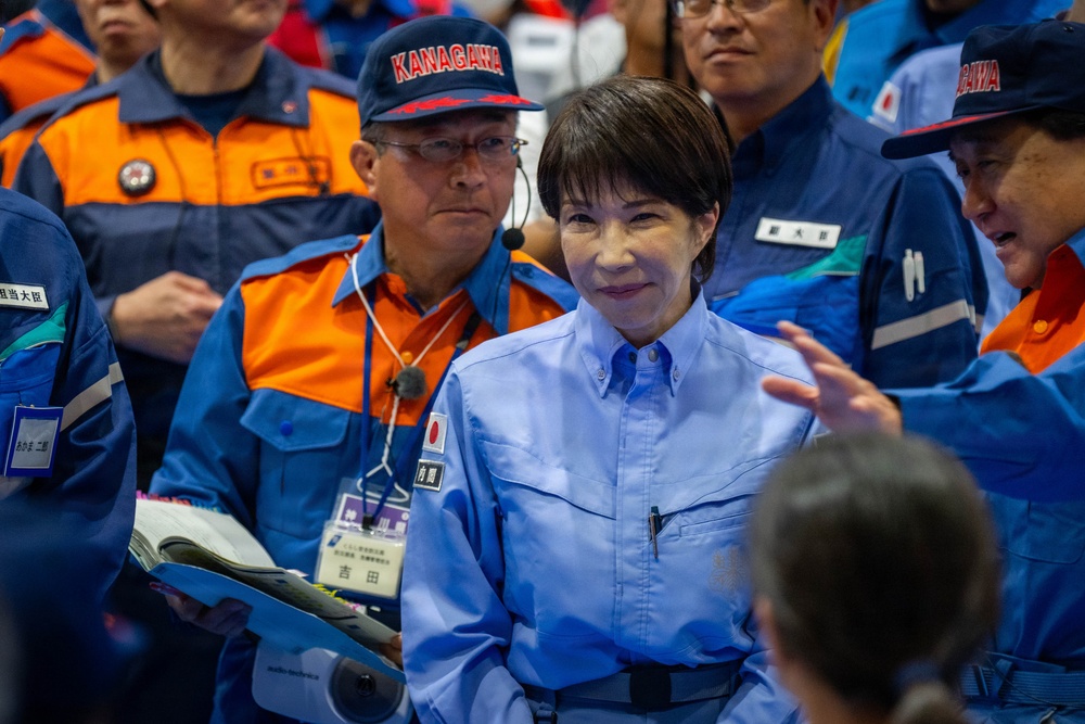 The image shows a group of individuals wearing official uniforms, likely at a Japanese emergency management or disaster response exercise. A central figure in a light blue uniform with a Japanese flag patch stands prominently among personnel wearing blue and orange uniforms marked 'KANAGAWA'. The setting appears to be an organized official event or drill.