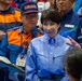 The image shows a group of individuals wearing official uniforms, likely at a Japanese emergency management or disaster response exercise. A central figure in a light blue uniform with a Japanese flag patch stands prominently among personnel wearing blue and orange uniforms marked 'KANAGAWA'. The setting appears to be an organized official event or drill.