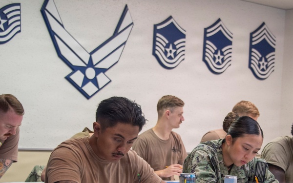 Military personnel from multiple branches, including U.S. Air Force and U.S. Navy, are seated at desks in a classroom setting, appearing to take a written exam or complete paperwork. The room is decorated with U.S. Air Force enlisted rank insignia and the Air Force symbol on the wall. Several energy drinks and water bottles are visible on the desks, suggesting an extended study or testing session.