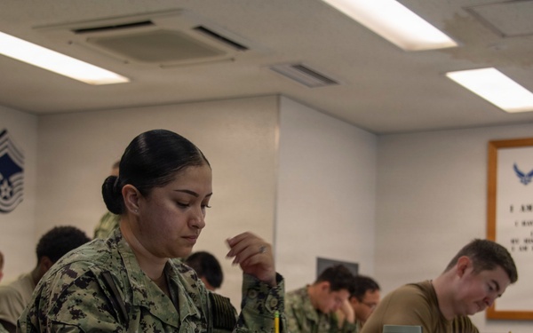 Military personnel from multiple branches are seated at desks in a classroom setting, appearing to take a written examination or complete paperwork. The room is decorated with U.S. Air Force insignia, rank chevrons, and a framed 'Airman's Creed' poster on the wall. The foreground features a female Navy sailor with the name tape 'ERDOMO' writing with a pencil.