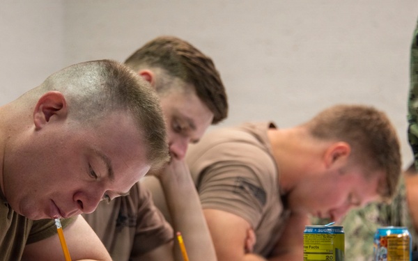 Several military personnel in uniform t-shirts are seated at a white table, writing with pencils on documents or test papers. A person in full camouflage uniform stands to the right, appearing to oversee the activity. Energy drink cans are visible on the table alongside papers and writing instruments.