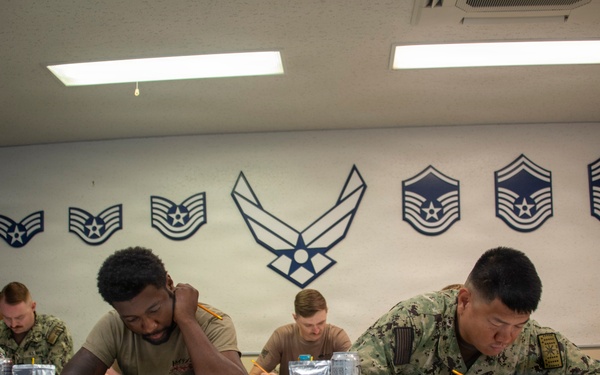 Military personnel in camouflage uniforms are seated at tables in a classroom or testing environment, writing on papers or documents. The room is decorated with U.S. Air Force enlisted rank insignia displayed on the wall, along with the central Air Force symbol (wings). The setting appears to be a formal military education or examination session.