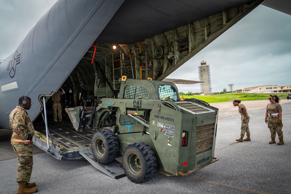 Military personnel are loading or unloading a Caterpillar skid-steer loader (marked USN 45-05717) onto or off a large military transport aircraft via a rear cargo ramp. The operation is taking place on an airfield tarmac, with a control tower visible in the background. Personnel in MultiCam/OCP uniforms are guiding the equipment during the operation.