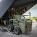 Military personnel are loading or unloading a Caterpillar skid-steer loader (marked USN 45-05717) onto or off a large military transport aircraft via a rear cargo ramp. The operation is taking place on an airfield tarmac, with a control tower visible in the background. Personnel in MultiCam/OCP uniforms are guiding the equipment during the operation.