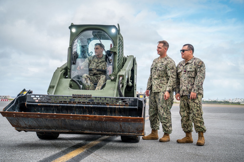 Three military personnel in U.S. Navy NWU Type III (woodland camouflage) uniforms are gathered on what appears to be a tarmac or airfield surface. One sailor is operating a military olive-drab skid steer loader (compact track loader) with a front bucket attachment, while two others stand nearby conversing. The setting appears to be an outdoor military installation with an overcast sky in the background.