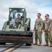 Three military personnel in U.S. Navy NWU Type III (woodland camouflage) uniforms are gathered on what appears to be a tarmac or airfield surface. One sailor is operating a military olive-drab skid steer loader (compact track loader) with a front bucket attachment, while two others stand nearby conversing. The setting appears to be an outdoor military installation with an overcast sky in the background.
