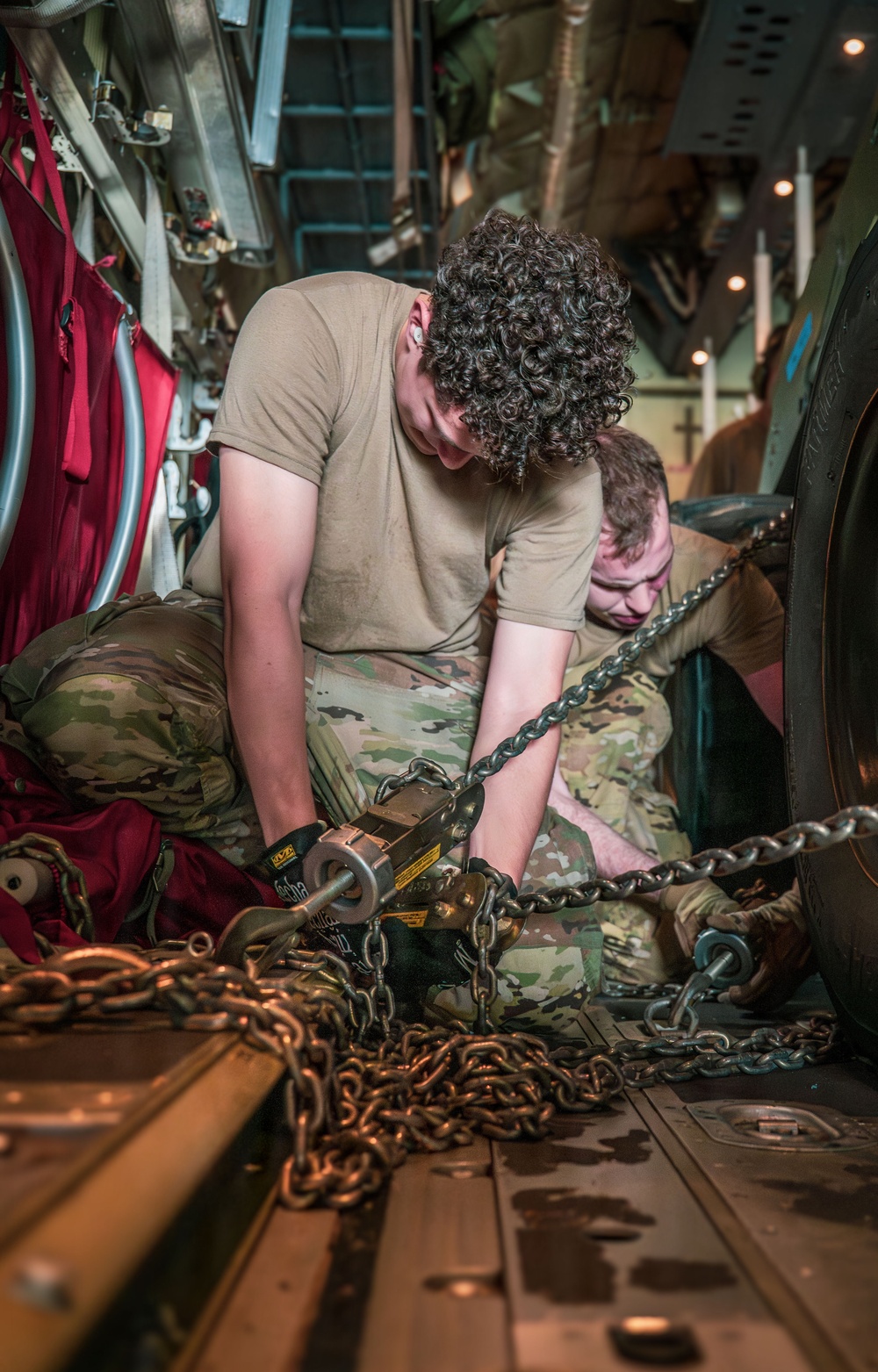 Two military personnel in OCP (Operational Camouflage Pattern) uniforms are working inside the cargo hold of a military aircraft, securing cargo with heavy chains and a chain binder/load binder tool. They are kneeling on the aircraft's cargo floor, which features standard tie-down rails, while working with a large pile of heavy-duty chains. The scene depicts standard cargo securing operations typical of military airlift missions.