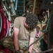 Two military personnel in OCP (Operational Camouflage Pattern) uniforms are working inside the cargo hold of a military aircraft, securing cargo with heavy chains and a chain binder/load binder tool. They are kneeling on the aircraft's cargo floor, which features standard tie-down rails, while working with a large pile of heavy-duty chains. The scene depicts standard cargo securing operations typical of military airlift missions.
