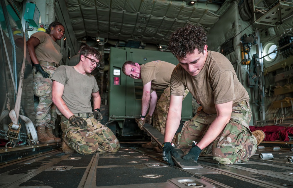 Four military personnel in U.S. Army/Air Force OCP (Operational Camouflage Pattern) uniforms are working inside the cargo hold of a military transport aircraft, likely a C-130 or similar airlifter. They appear to be securing or configuring cargo tie-down fittings and floor tracks. The scene depicts a loading or pre-mission preparation operation.