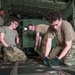 Four military personnel in U.S. Army/Air Force OCP (Operational Camouflage Pattern) uniforms are working inside the cargo hold of a military transport aircraft, likely a C-130 or similar airlifter. They appear to be securing or configuring cargo tie-down fittings and floor tracks. The scene depicts a loading or pre-mission preparation operation.