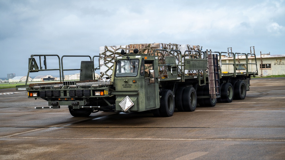 A US Air Force military cargo truck is operating on what appears to be an airfield tarmac, transporting palletized cargo secured with netting. The olive-drab vehicle is marked 'US AIR FORCE' and identifier '04E00025', and is towing an additional flatbed trailer. The setting appears to be an active military air base under overcast skies.