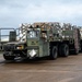 A US Air Force military cargo truck is operating on what appears to be an airfield tarmac, transporting palletized cargo secured with netting. The olive-drab vehicle is marked 'US AIR FORCE' and identifier '04E00025', and is towing an additional flatbed trailer. The setting appears to be an active military air base under overcast skies.